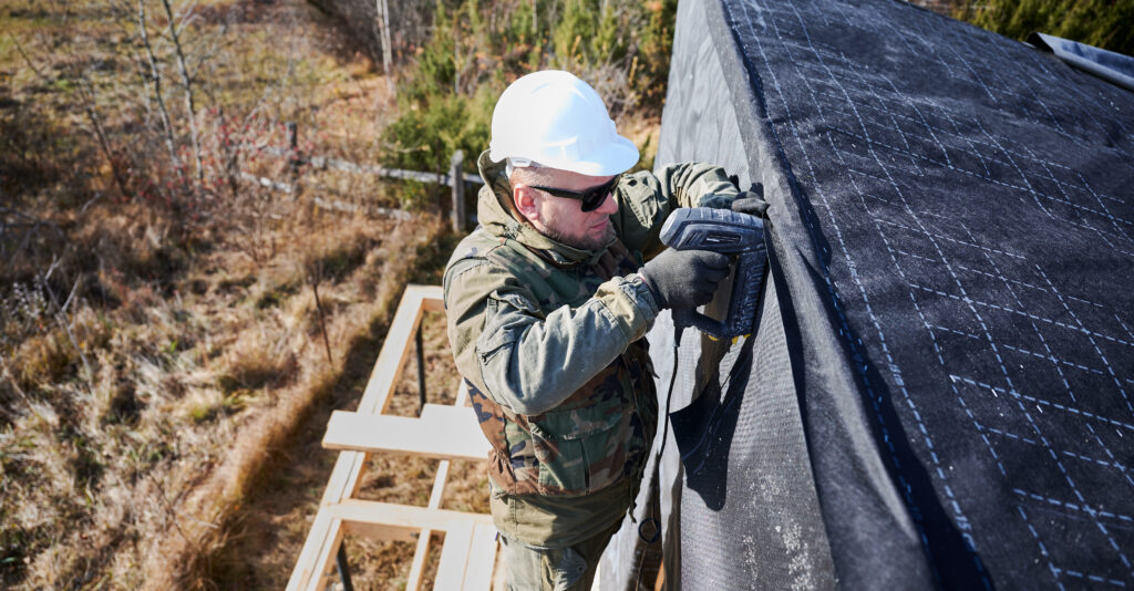 Male builder installing waterproof membrane on wall of future wooden frame house.