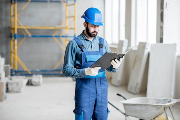 Builder or foreman in working uniform expertising the structure standing with folder at the construction site indoors